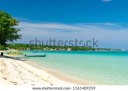 Sunny day along the Seven Mile Beach in tropical Negril, Jamaica. Tour boats await passengers and caucasian tourists in the water at a distance. Caribbean Jamaican summer beach vacation. Royalty-Free Stock Photo #1161489259