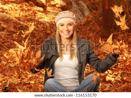Picture of happy blonde girl having fun in autumn woods, pretty woman throwing up dry autumnal leaves in the forest, beautiful female sitting down on ground covered golden foliage