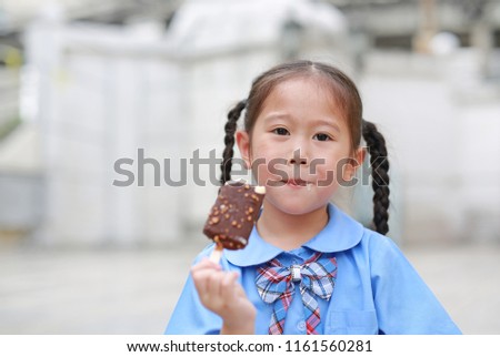 Happy little Asian child girl in school uniform enjoy eating chocolate vanilla ice cream outdoors.