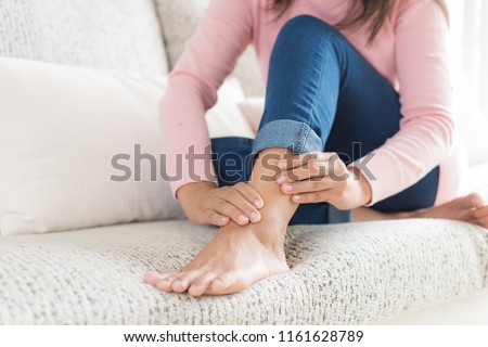 Closeup woman sitting on sofa holds her ankle injury, feeling pain. Health care and medical concept. Royalty-Free Stock Photo #1161628789