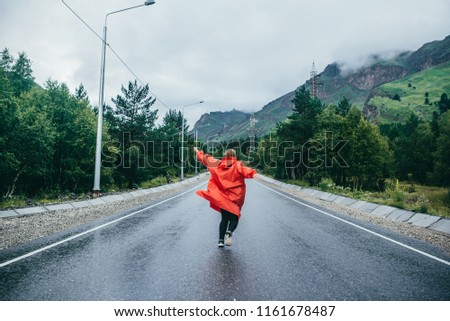 Woman tourist runs in raincoat or ashes on wet, paved road after rain in mountains