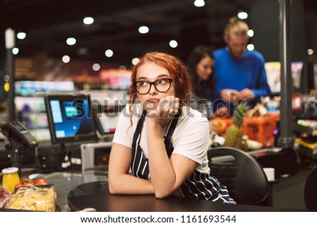 Young boring cashier in eyeglasses and striped apron dreamily looking aside working in modern supermarket with customers on background