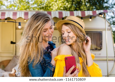 Cheerful girls do selfie on a smartphone on the back of a van in a camping. Road trip with best friends. Adventure, freedom, youth