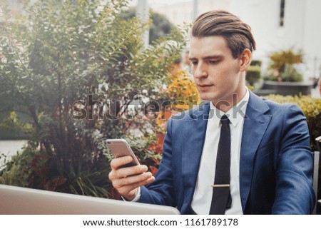Young attractive pensive businessman holding mobile phone in hand and sitting next to green plant.