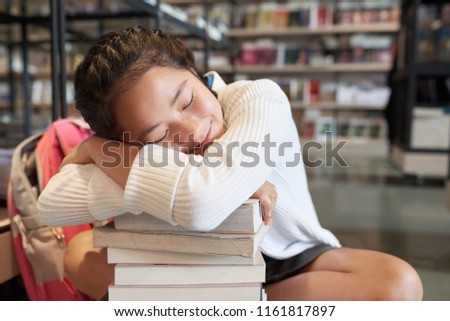 Young student girl with closed eyes sitting leaning head on arms on pile of books smiling on blurred bookshelves background