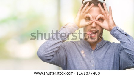 Handsome young elegant man wearing glasses doing ok gesture like binoculars sticking tongue out, eyes looking through fingers. Crazy expression.