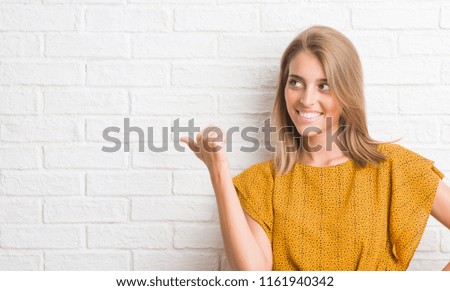 Beautiful young woman over white brick wall pointing and showing with thumb up to the side with happy face smiling