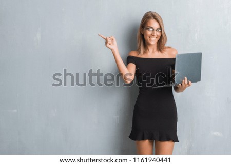 Beautiful young business woman over grunge grey wall using computer laptop very happy pointing with hand and finger to the side