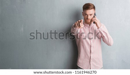Young redhead man over grey grunge wall talking on the phone pointing with finger to the camera and to you, hand sign, positive and confident gesture from the front