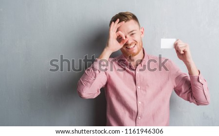 Young redhead man over grey grunge wall holding blank visit card with happy face smiling doing ok sign with hand on eye looking through fingers