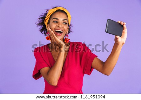 Joyful african woman in dress making selfie on smartphone while touching her cheek over purple background