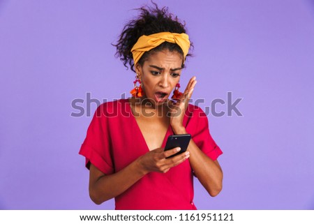 Shocked african woman in dress using smartphone with open mouth over purple background