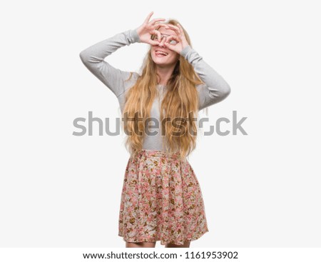 Blonde teenager woman wearing flowers skirt doing ok gesture like binoculars sticking tongue out, eyes looking through fingers. Crazy expression.