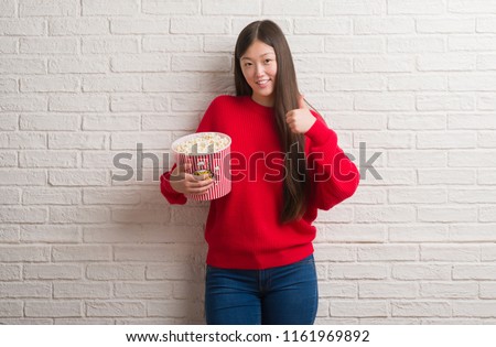 Young Chinese woman over brick wall eating pop corn happy with big smile doing ok sign, thumb up with fingers, excellent sign