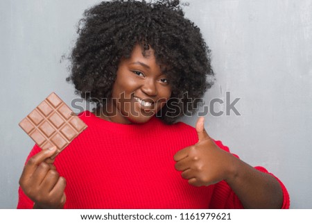 Young african american woman over grey grunge wall eating a chocolate bar happy with big smile doing ok sign, thumb up with fingers, excellent sign