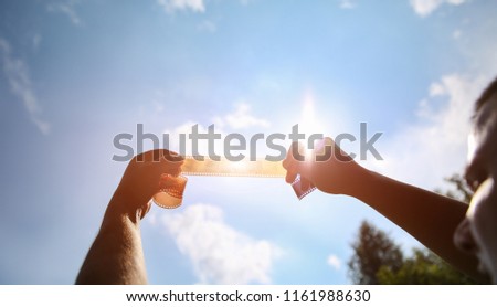 Man is holding film strip in his hands against the sunlight and sky. Guy remembering his childhood memories in the nature. Retro concept on the photo tape.