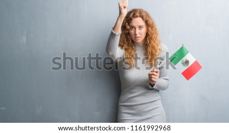 Young redhead woman over grey grunge wall holding flag of Mexico annoyed and frustrated shouting with anger, crazy and yelling with raised hand, anger concept