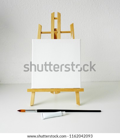 Wooden easel, empty white linen canvas, brush for drawing and tube of white paint 