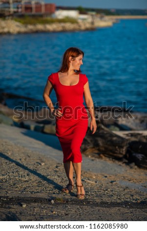 A red-haired young woman in a long red dress is standing on a concrete breakwater on the beach. A young beautiful woman in a red dress by the figure stands on a pier by the pond.