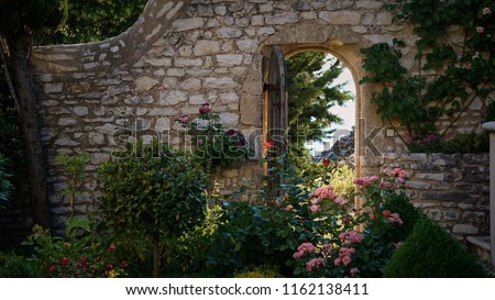 Rear view of a fairy like rose garden with an ancient stone wall and an open wooden door                          