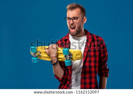 Charismatic cheerful young bearded man dressed in checkered shirt, white T-shirt and glasses, with yellow skateboard in his hands. Studio shot with blue background