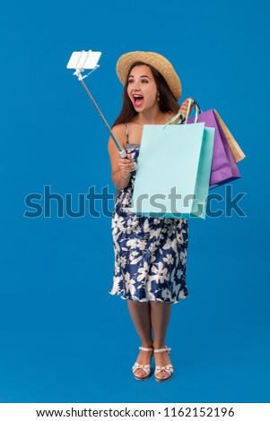 Young woman in casual clothes with shopping bags using selfie stick to take a self portrait on blue studio background with copy space