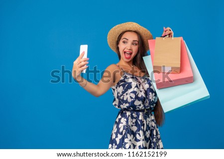 Young woman in casual clothes with shopping bags using selfie stick to take a self portrait on blue studio background with copy space