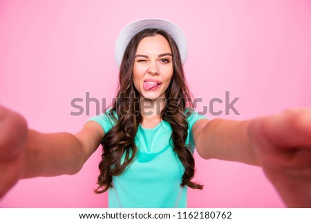 Pretty young girl in a white hat make selfie on front camera smartphone wink and show thoung out isolated on shine pink background