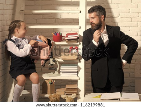 Family stands next to shelf with school supplies. Schoolgirl and dad with smiling faces hold pink schoolbag. Back to school and fathers day concept. Girl and man in classroom on white brick background