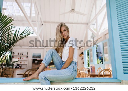 Inspired barefooted woman with pretty smile posing in cafe. Photo of cute blonde female model with excited face expression sitting on window sill.