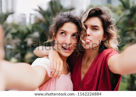 Carefree fair-haired lady making selfie during walk with friend. Blissful brunette girl embracing her sister on blur nature background.