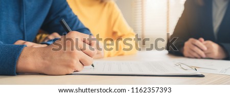 Happy young Asian couple and realtor agent. Cheerful young man signing some documents while sitting at desk together with his wife. Signing good condition contract. Panoramic banner background.