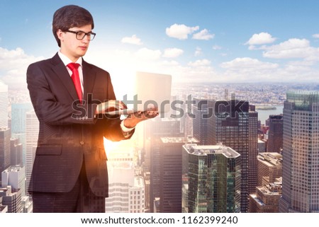 Handsome young businessman in glasses holding a laptop standing over modern city background on a sunny day. Toned image double exposure mock up