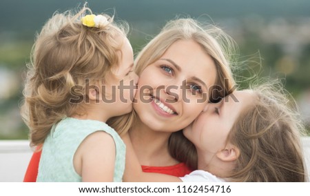 Daughters kiss mother on natural landscape. Mothers day concept. Woman and girls sit on bench. Happy childhood, family, love. Summer vacation, leisure, activity, lifestyle