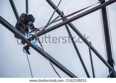 A working man is an engineer and welder in a construction overall, a welding mask is cooking metal and is sitting on a metal structure at an altitude against the blue sky