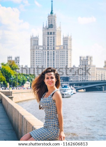 A girl in a blue dress in a cage with long hair stands and smiles on the background of a Stalin skyscraper in Moscow on a Sunny day with a beautiful sky near the river