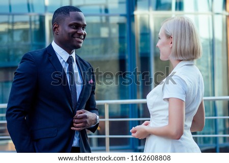 handsome African American businessman in a stylish black suit talking with attractive blonde woman business lady on the street background office skyscraper