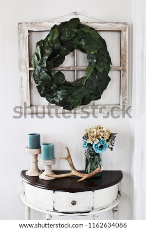 Old farmhouse window decorated with a homemade Magnolia leaf wreath hung on an interior wall over rustic half moon table with candles and flowers decor.
