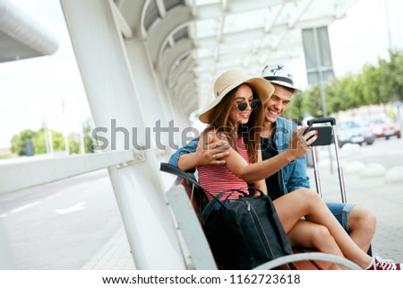 Travel. Couple Taking Photos On Phone Sitting At Bus Stop Near Airport Outdoors. Young People Traveling In Summer. High Resolution
