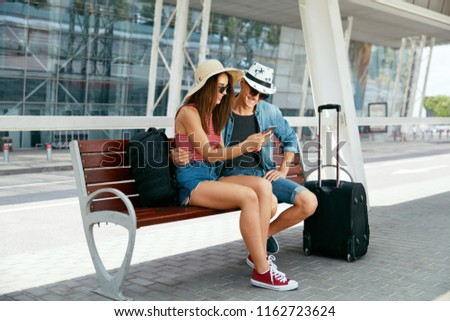 Couple Using Phone Making Video Call Or Taking Photos Sitting At Bus Stop With Suitcase Near Airport. High Resolution