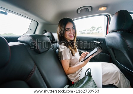 A young Japanese Asian woman sits and reads her e reader while sitting in the backseat of a car she booked via a ride hailing app. She is smiling happily as she reads and is driven to her destination. Royalty-Free Stock Photo #1162739953