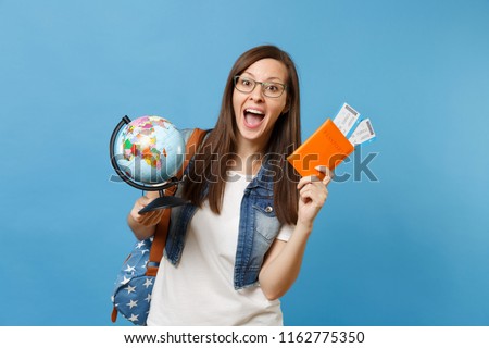 Young happy woman student in glasses with backpack holding world globe, passport, boarding pass tickets isolated on blue background. Education in university college abroad. Air travel flight concept Royalty-Free Stock Photo #1162775350