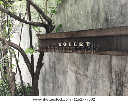 Toilet sign on a wooden board hanging on concrete wall beside branches of trees