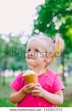 little funny girl blonde eating sweet blue ice cream in a waffle cup on a green summer background in the park. smeared her face and cheeks and laughs. Dressed in bright stylish clothes.
