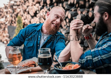 Arm wrestling. Bearded red-haired man having no strength for battling in arm wrestling his best friend