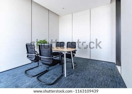 Part of ordinary office room decorated in modern style with meeting room,large windows with blinds,carpeting,ventilation,escape signs,white furniture (tables, shelves, drawers) and dark office chairs.