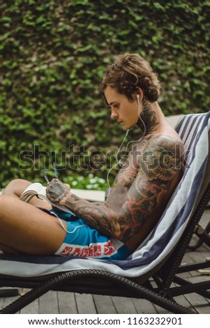 a young man in tattoos wearing headphones listens to music and draws a pencil in a notebook. Online training.