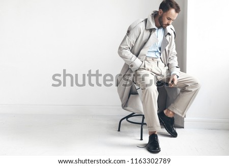 Fashion portrait of handsome  stylish man with dark beard and hair, weared in light trench coat, shirt, beige pants and black shoes. Man sits on the chair near white wall 