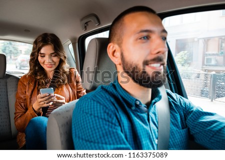 Smiling driver talking with female passenger. Woman using mobile phone in the background. Royalty-Free Stock Photo #1163375089