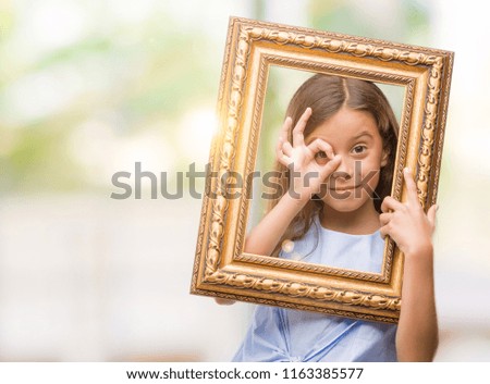Brunette hispanic girl holding vintage art frame with happy face smiling doing ok sign with hand on eye looking through fingers